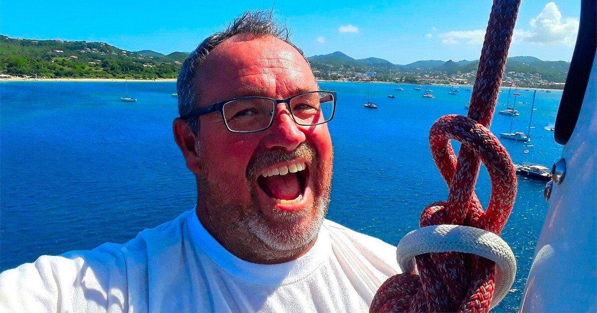Mark Haswell, captain of the Rapido 60 trimaran Ineffable, smiling on deck against a tropical bay backdrop.