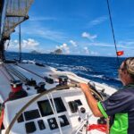 Person steering a Rapido trimaran under sail on open water, with wind in their hair and islands on the horizon.