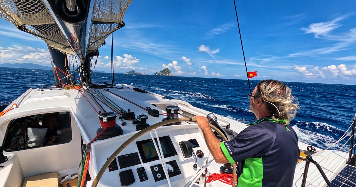 Person steering a Rapido trimaran under sail on open water, with wind in their hair and islands on the horizon.