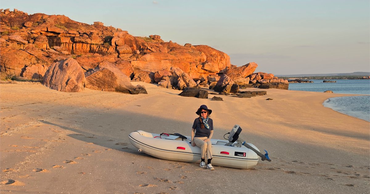 Merri sitting on a dinghy on a deserted beach at Jar Island, Kimberley, Western Australia, with dramatic red sandstone cliffs in the background.
